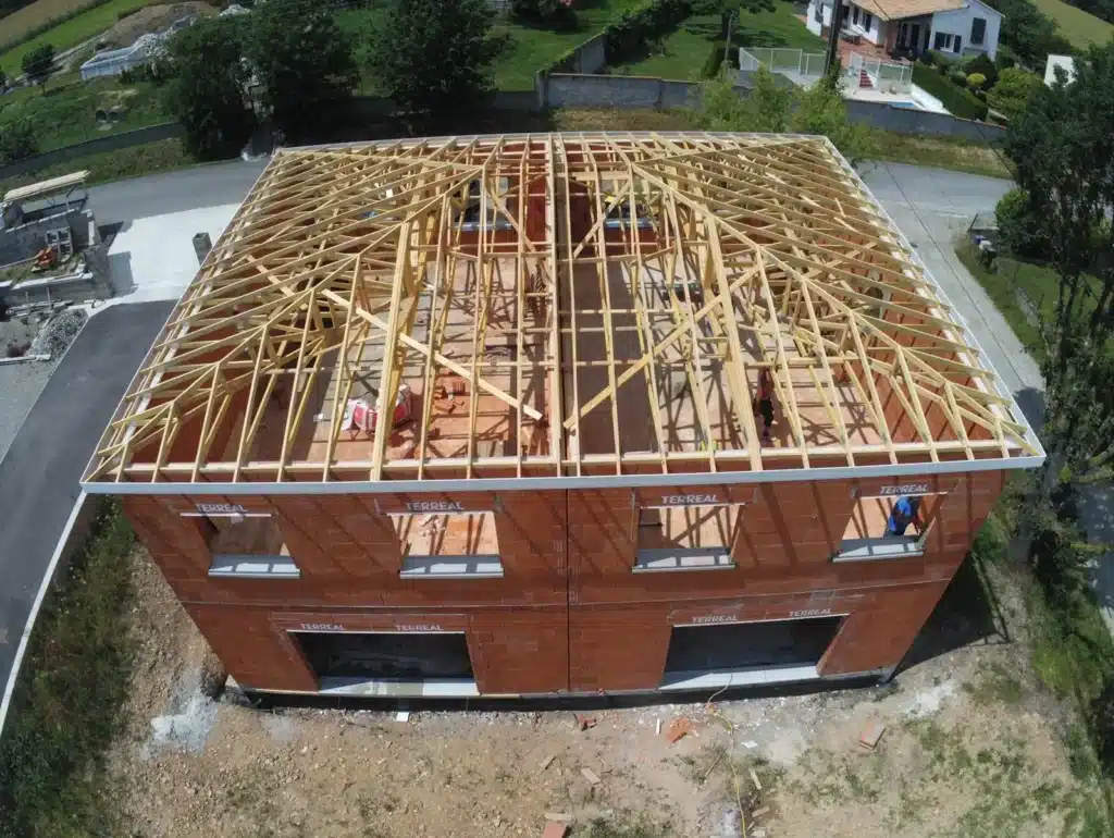 Vue aérienne d'une charpente industrielle à fermettes en cours d'installation sur une maison neuve en briques rouges, mettant en avant l'expertise d'un charpentier dans la construction de grandes structures.